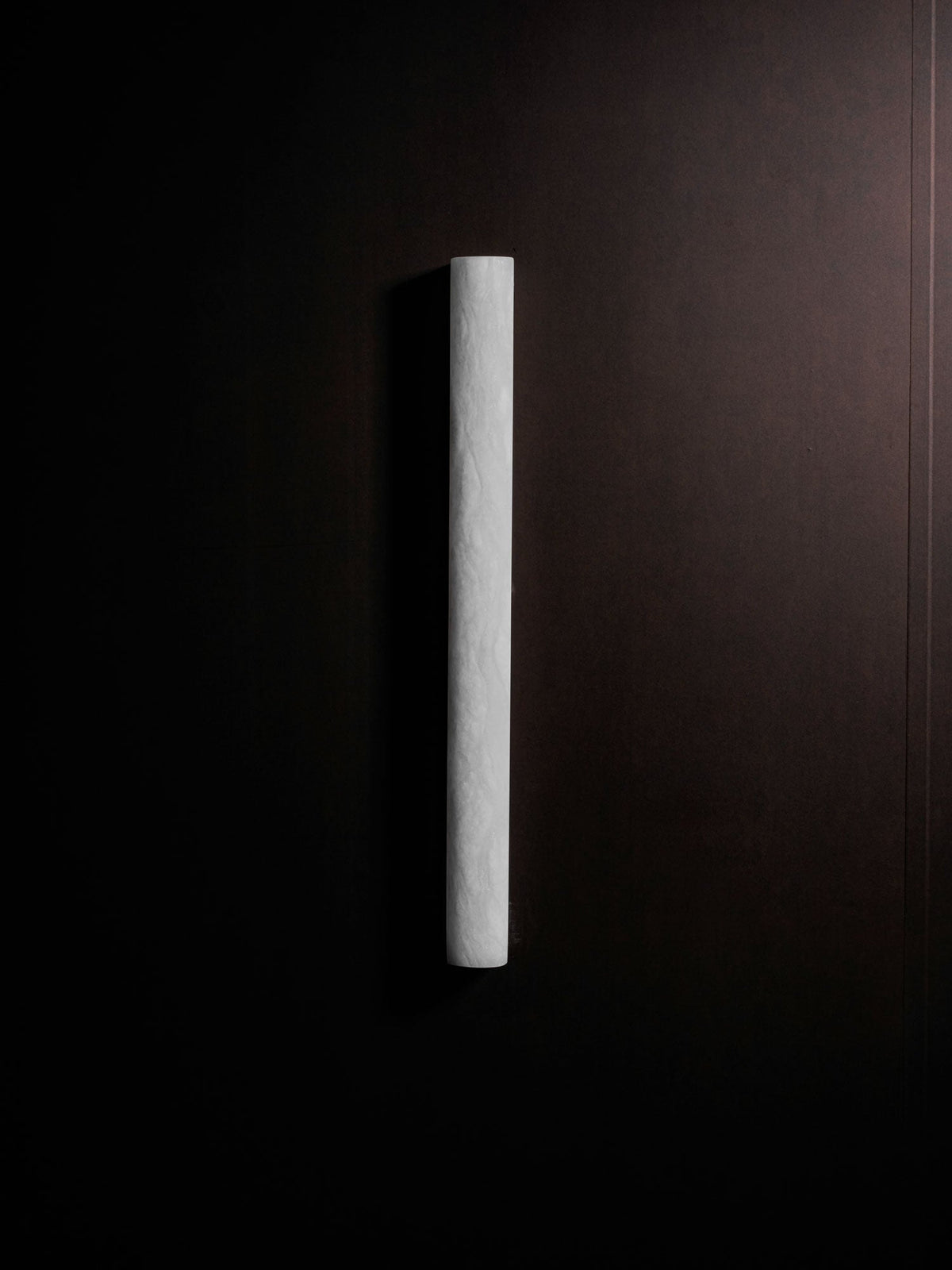 Unlit slim Clérachic sconce in pale stone on a dark brown wall, a narrow vertical bar with a soft, rough surface.