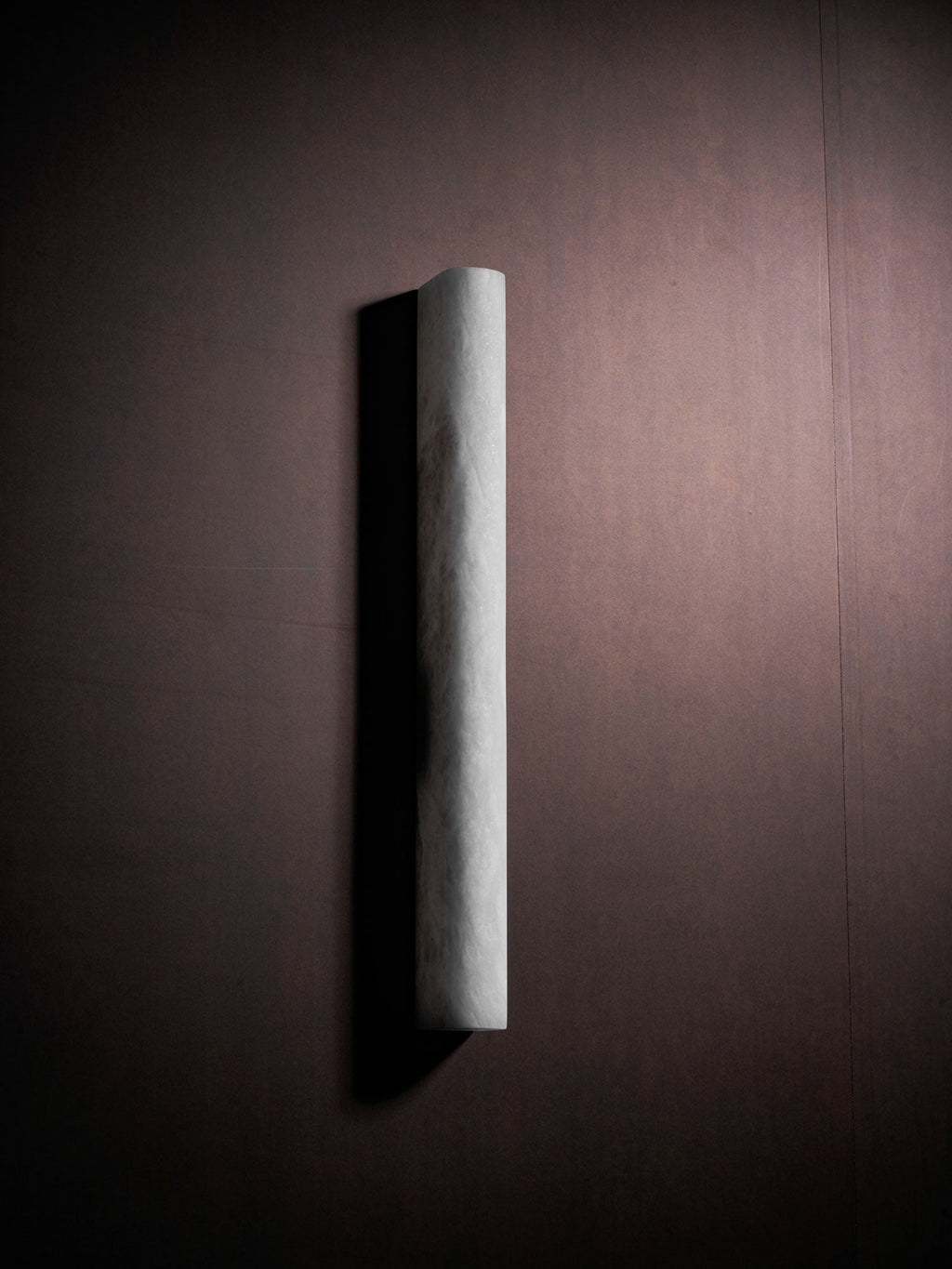 A Clérachic alabaster sconce, unlit, at an angle on a dark plum-brown wall, its pale curved face casting a shadow to the left.