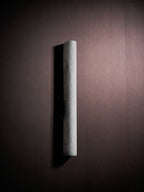 A Clérachic alabaster sconce, unlit, at an angle on a dark plum-brown wall, its pale curved face casting a shadow to the left.
