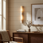 A Clérachic alabaster cylinder wall light glows on a panelled wall above a walnut sideboard, beside a framed abstract print and dried stems.