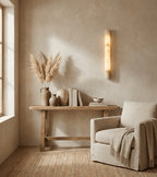 This Clérachic alabaster wall light glows beside a window over a rustic console holding pampas grass, vases and books, with a linen armchair.