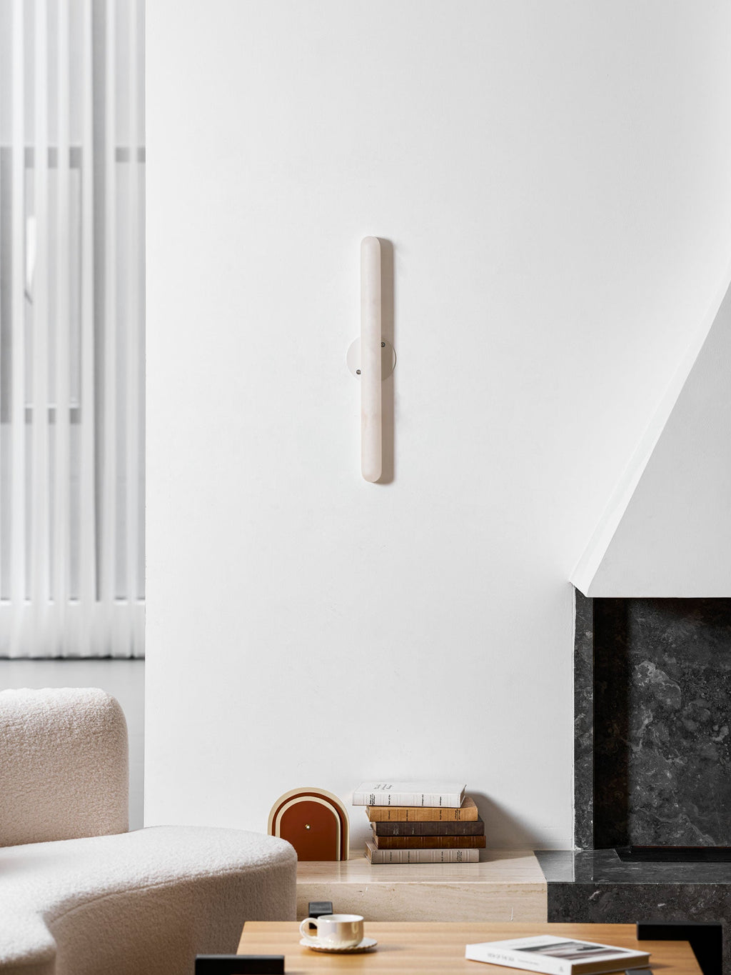 Unlit slim Clérachic alabaster wall light on a white wall above a boucle sofa, a coffee table and a dark stone fireplace.