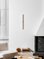 Unlit slim Clérachic alabaster wall light on a white wall above a boucle sofa, a coffee table and a dark stone fireplace.