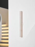 Close, angled view of the unlit slim Clérachic alabaster wall light on a white wall, with a blurred lit staircase at left.