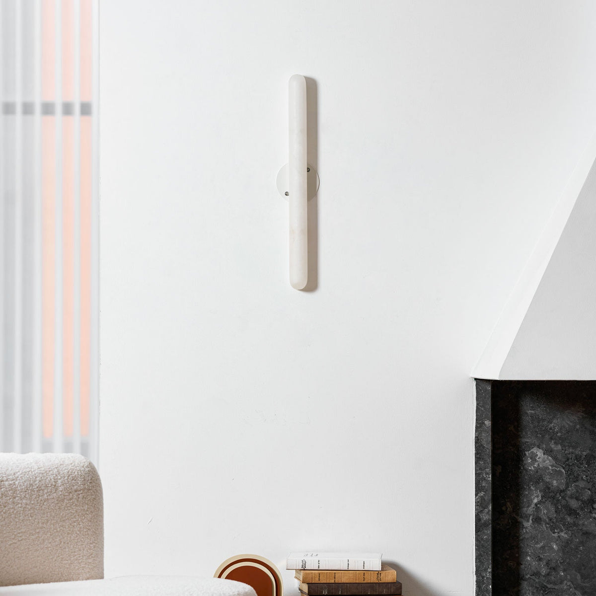 Unlit Clérachic alabaster wall light high on a white wall above a boucle sofa corner and a dark stone fireplace surround.