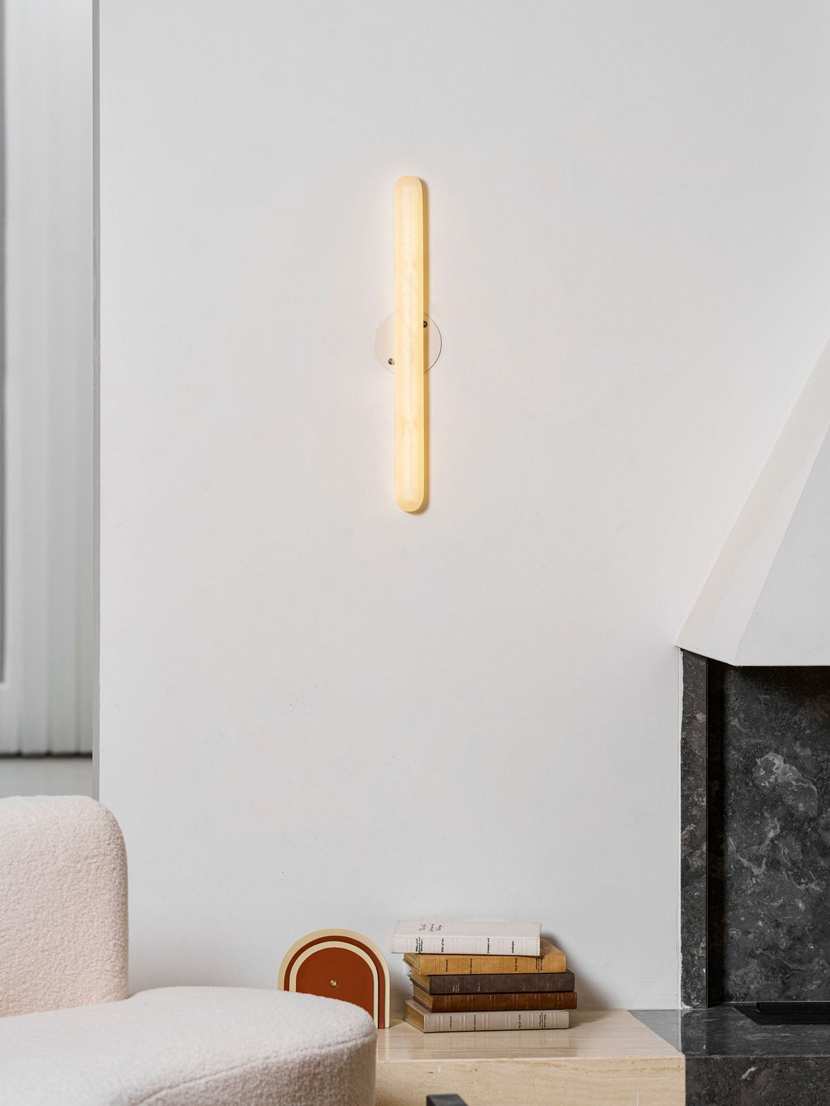 Clérachic alabaster wall light switched on, glowing a warm yellow on a white wall above a boucle sofa and a stack of books.