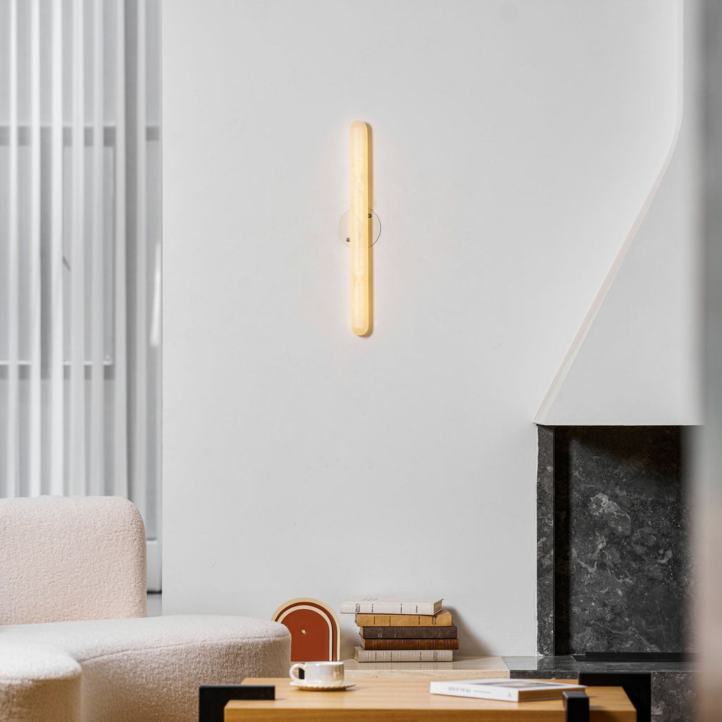 Lit slim Clérachic alabaster wall light on a white wall, with a boucle sofa, a coffee table and a dark stone fireplace below.