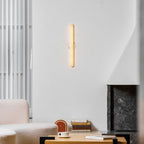 Lit slim Clérachic alabaster wall light on a white wall, with a boucle sofa, a coffee table and a dark stone fireplace below.