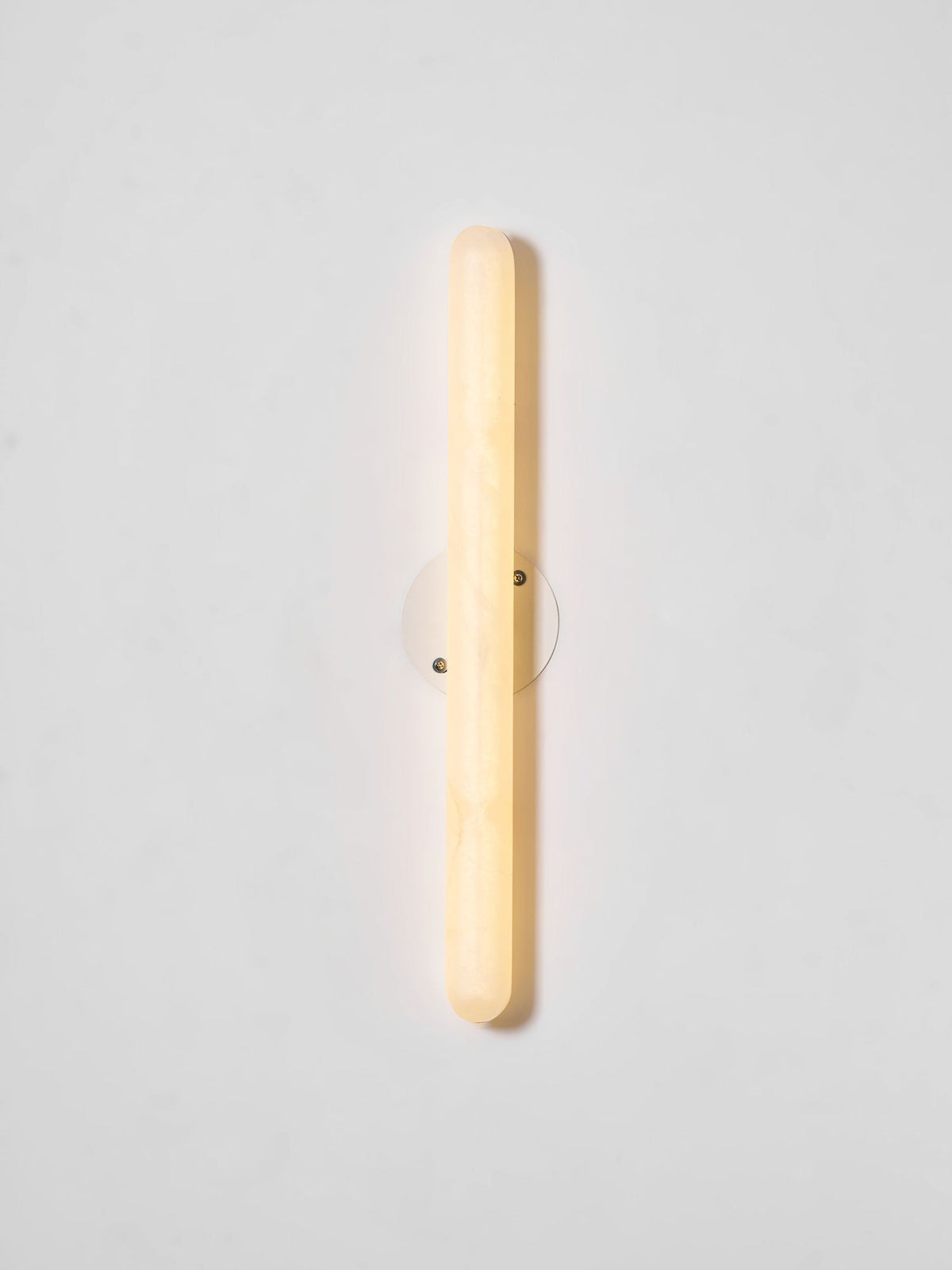 Lit Clérachic alabaster wall light on a pale wall, with the stone cylinder glowing warm in front of its small round backplate.