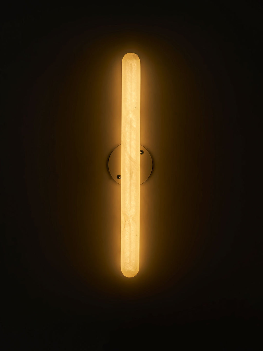 This Clérachic alabaster wall light glowing amber against a dark backdrop, with a small round backplate behind the stone cylinder.