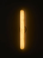 Close-framed Clérachic alabaster wall light lit in a dark room, the translucent cylinder glowing gold from end to end.