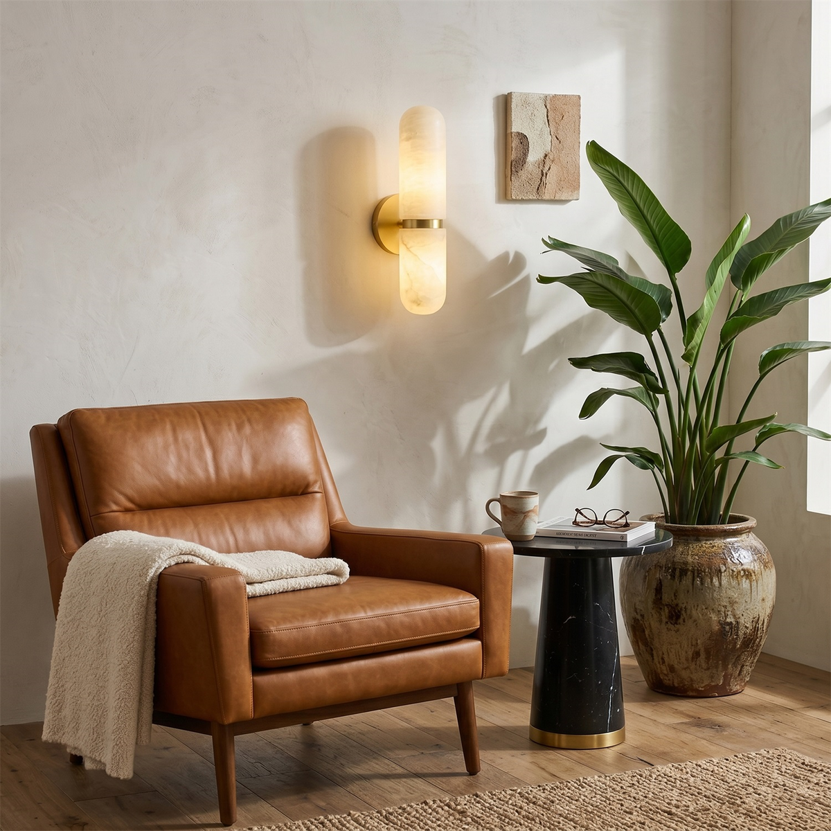 Lit Clérachic alabaster wall light above a tan leather armchair with a cream throw, beside a black side table and a large plant.
