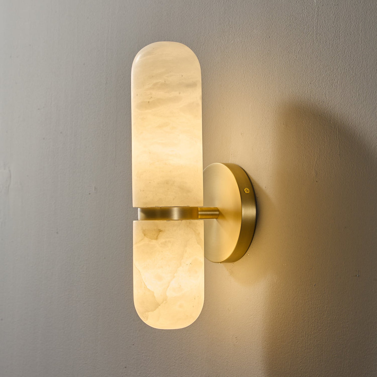 Lit Clérachic alabaster wall light on a grey wall, its veined stone shade glowing warm above and below the brass band.