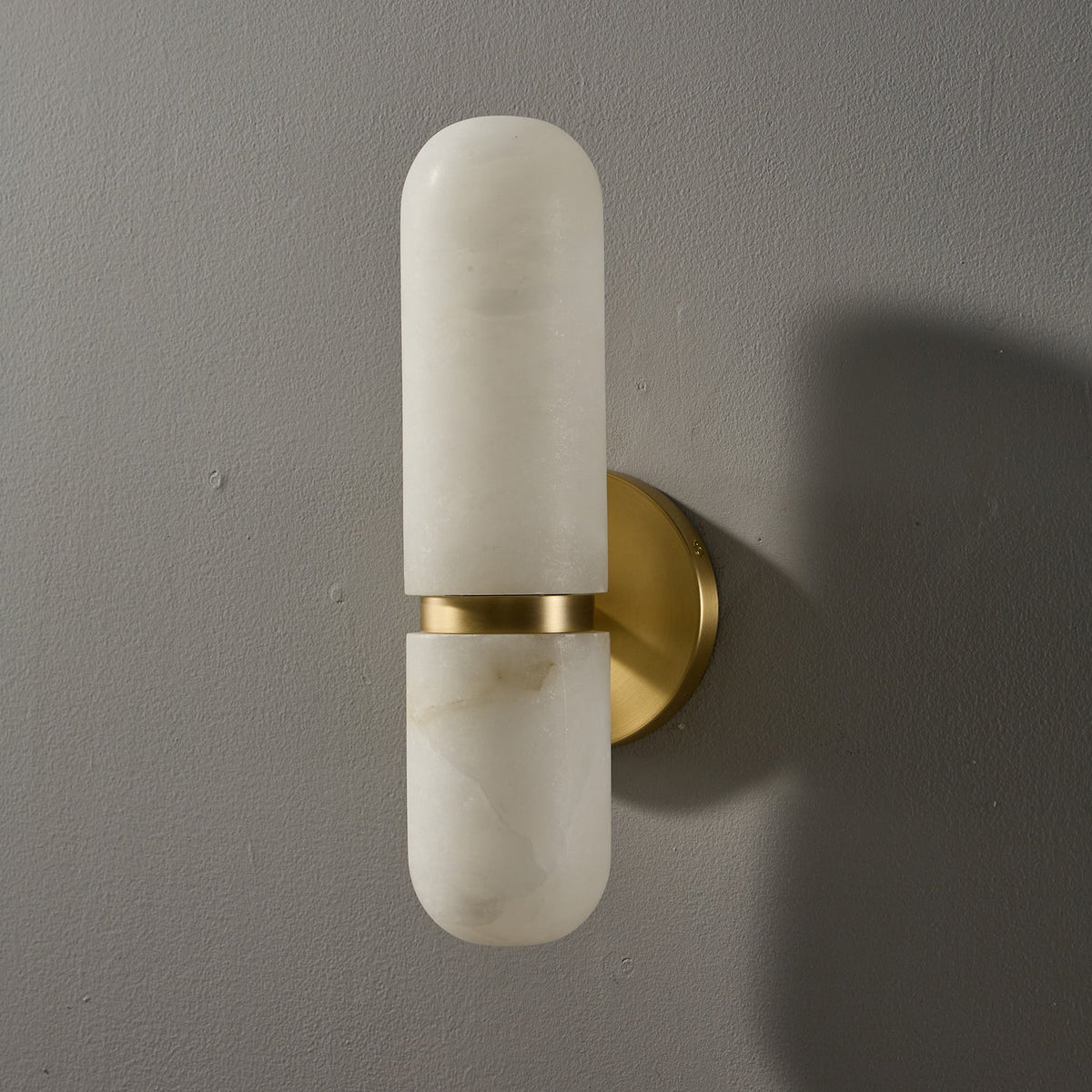 Clérachic alabaster wall light, unlit, on a grey wall, with a brass band and round brass backplate casting a shadow.
