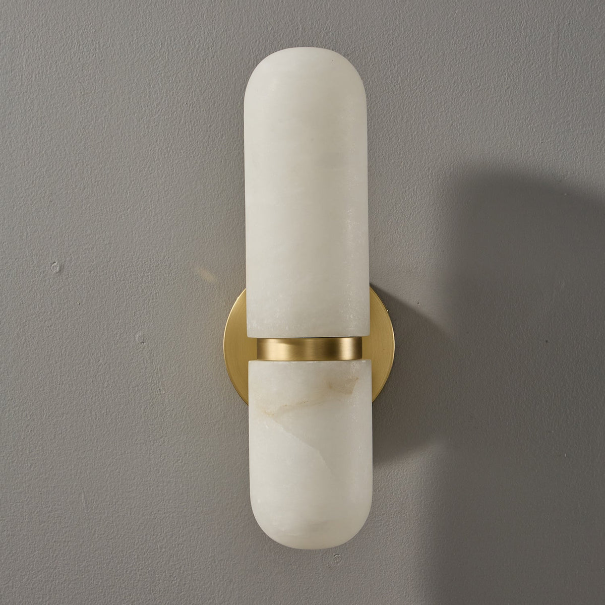 Front view of the unlit Clérachic alabaster wall light, with a pill-shaped shade split by a brass band over the round backplate.