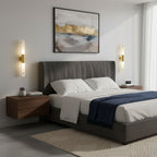 Two Clérachic gold-and-white wall lights flank a grey upholstered bed, with a navy throw, a floating nightstand and abstract art above.