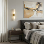 A Clérachic gold-and-white wall light glowing beside a grey bed with layered cushions, under a large abstract painting.
