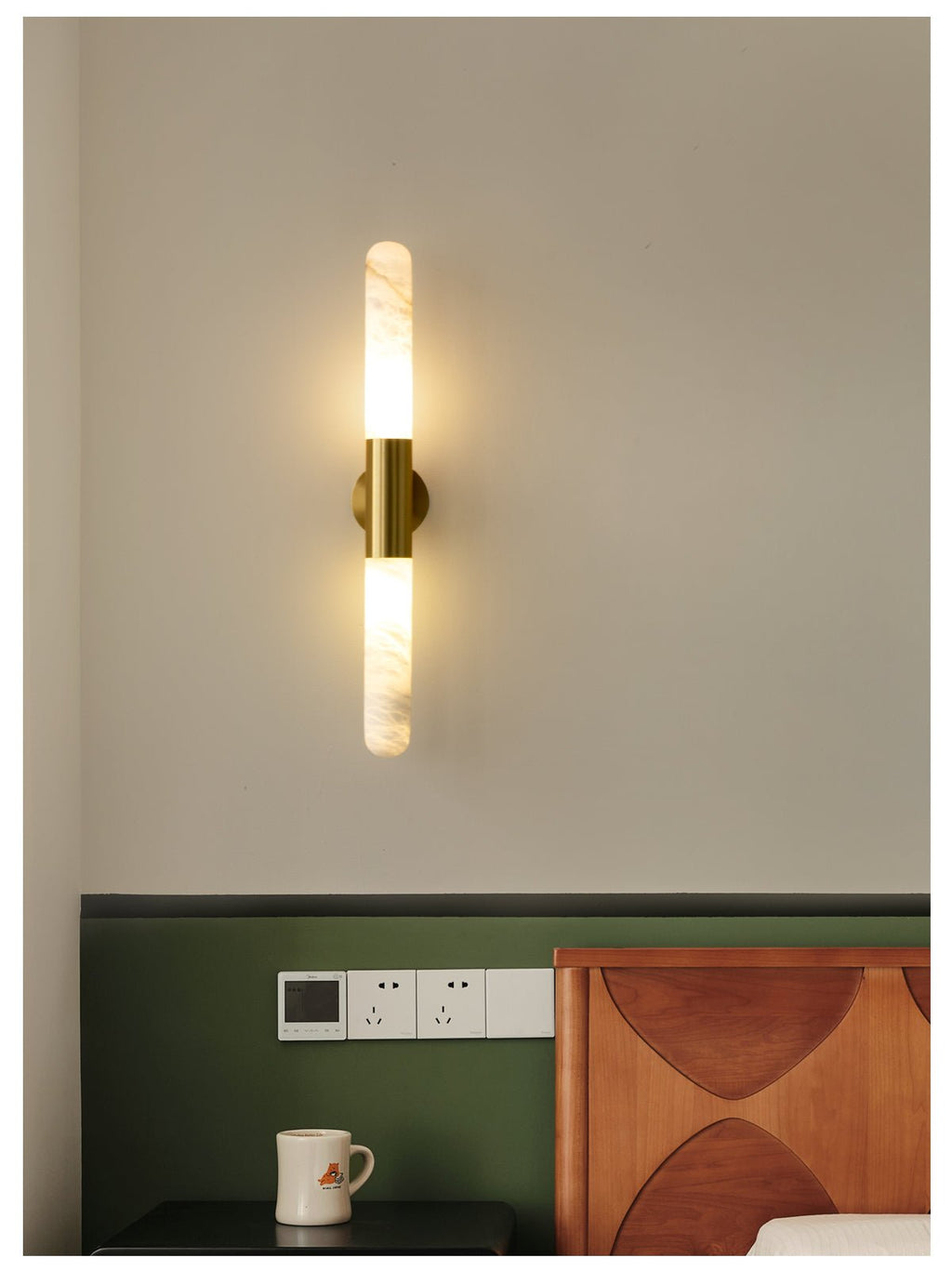 Lit Clérachic gold-and-white wall sconce above a green wall panel, with a socket plate and a carved wooden headboard below.