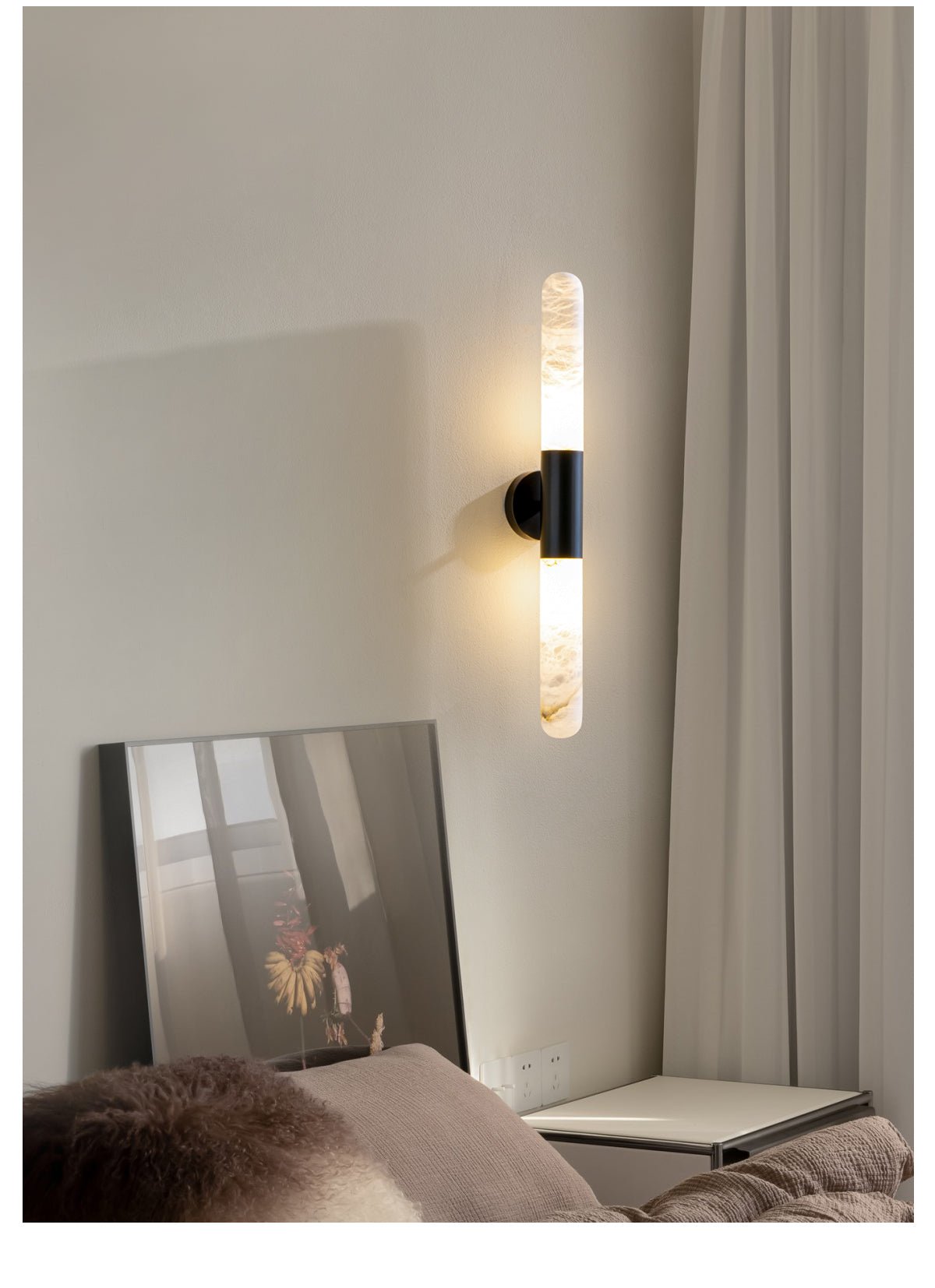 A black-and-white Clérachic wall light glowing beside a sheer curtain, above a leaning mirror and a bedside surface.