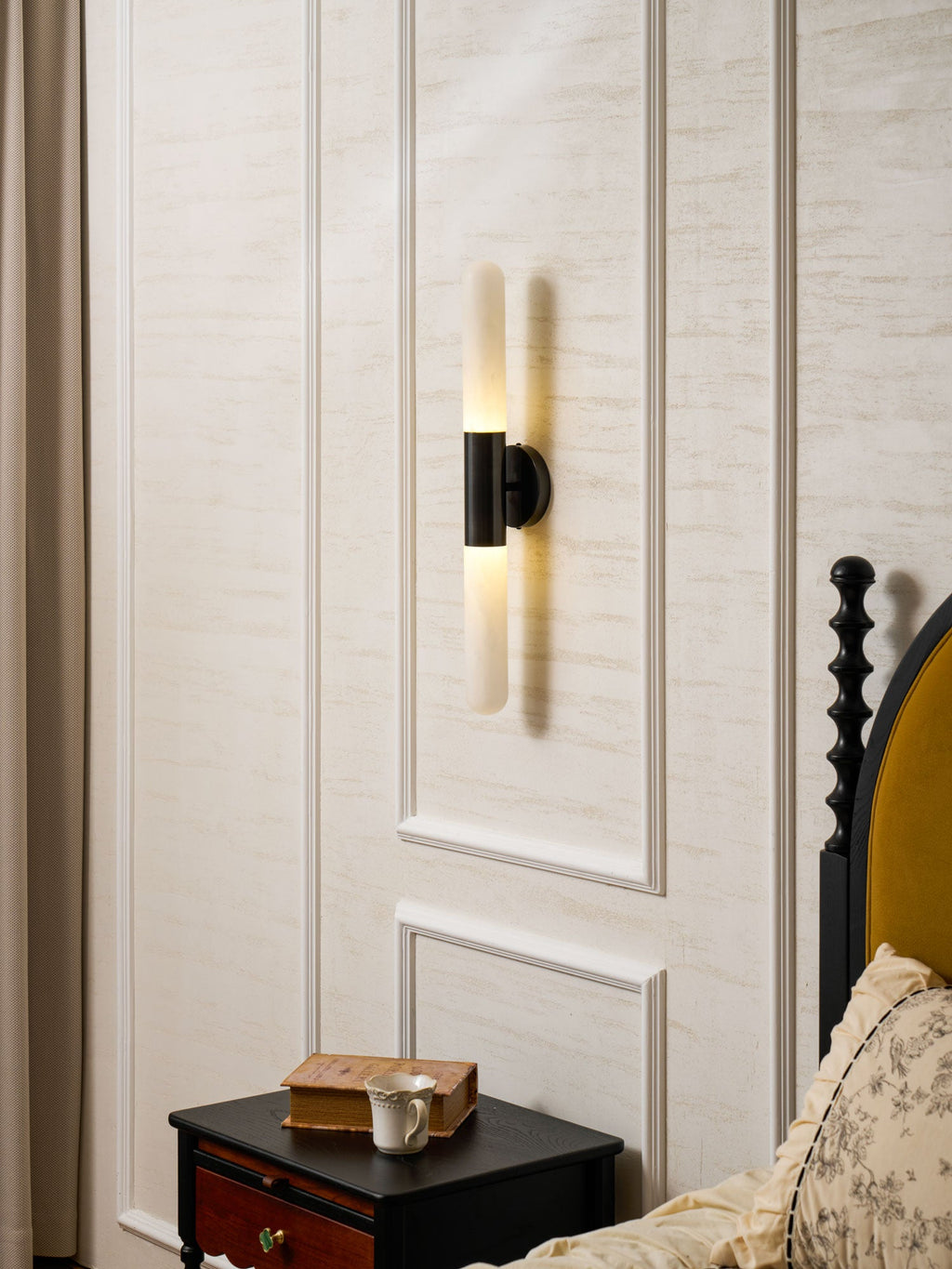 Lit Clérachic black-and-white wall light on a panelled cream wall, above a dark nightstand with a book and cup, beside a bed.
