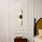 This Clérachic wall light glowing softly in the centre of a moulded wall panel, with a dark bed frame and gold headboard on the right.
