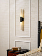 An angled view of a lit Clérachic black-and-white wall sconce on panelled walls, with a dark bedside table and floral bedding nearby.