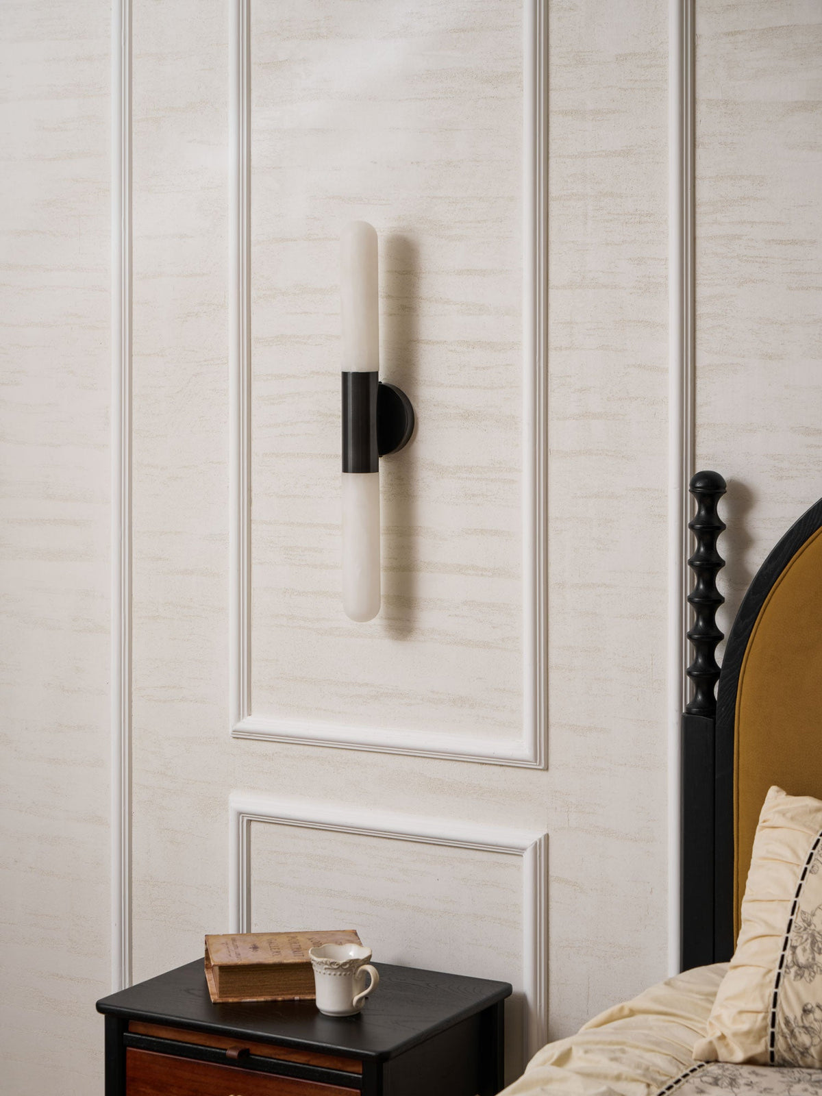 Unlit Clérachic wall light with a black centre band and white alabaster ends, mounted on a panelled wall above a bedside table.