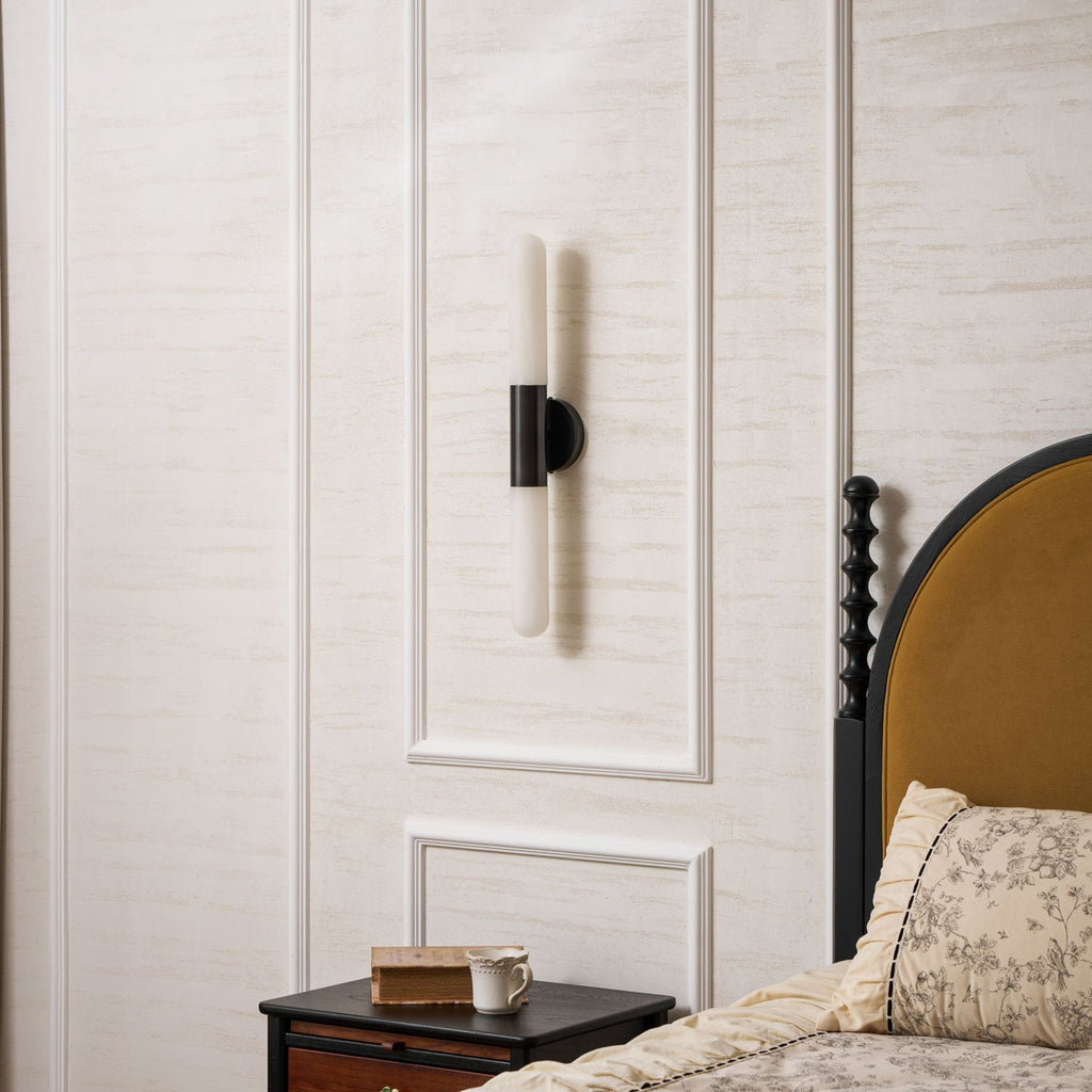 A Clérachic black-and-white wall light, switched off, centred on a wall panel beside a dark bed frame with an ochre headboard.