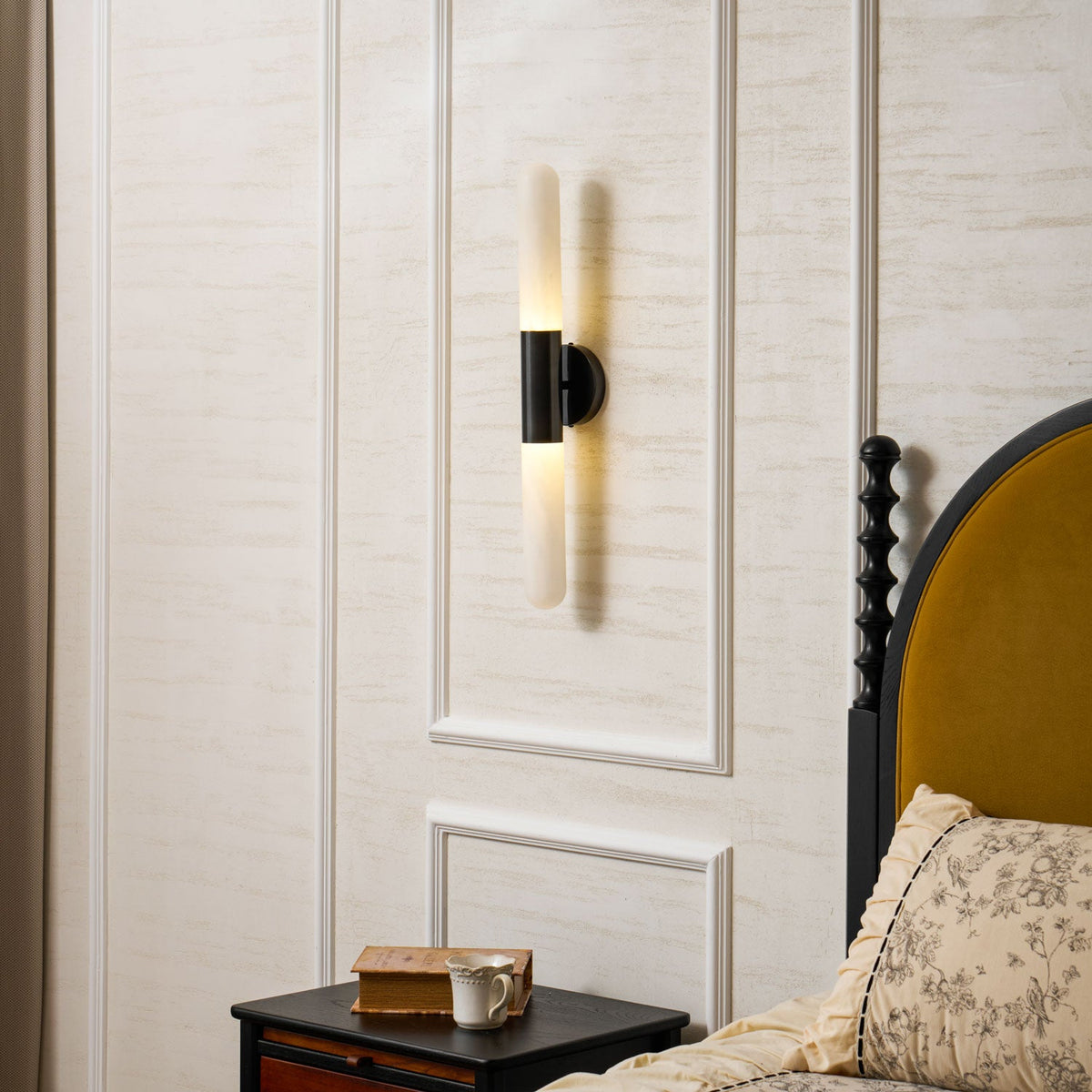 Lit Clérachic black-and-white wall light on a panelled wall, a pool of light rising and falling above a nightstand and floral bedding.