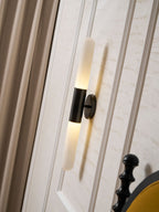Tilted view of a lit Clérachic black-and-white wall sconce on a panelled wall, with curtains to the left and a bed post below.