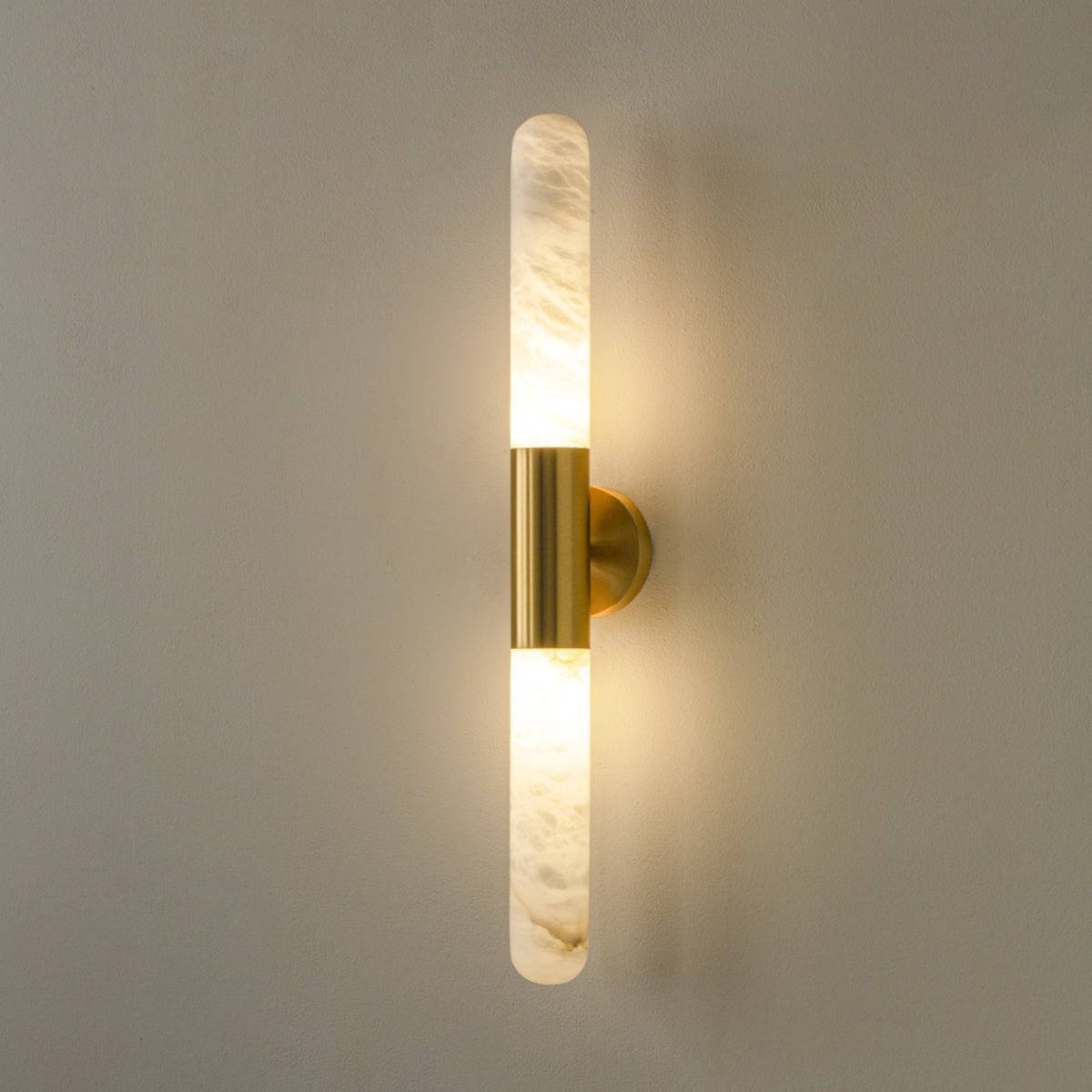 Close-up of the veined alabaster and the brass centre band of a lit Clérachic wall light on a grey-green wall.
