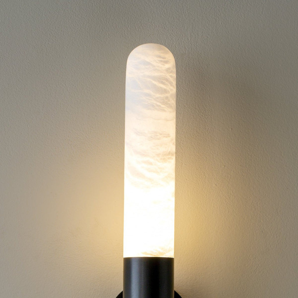 Close view of one lit alabaster tube of this Clérachic wall light, with a black collar and warm light at its base.