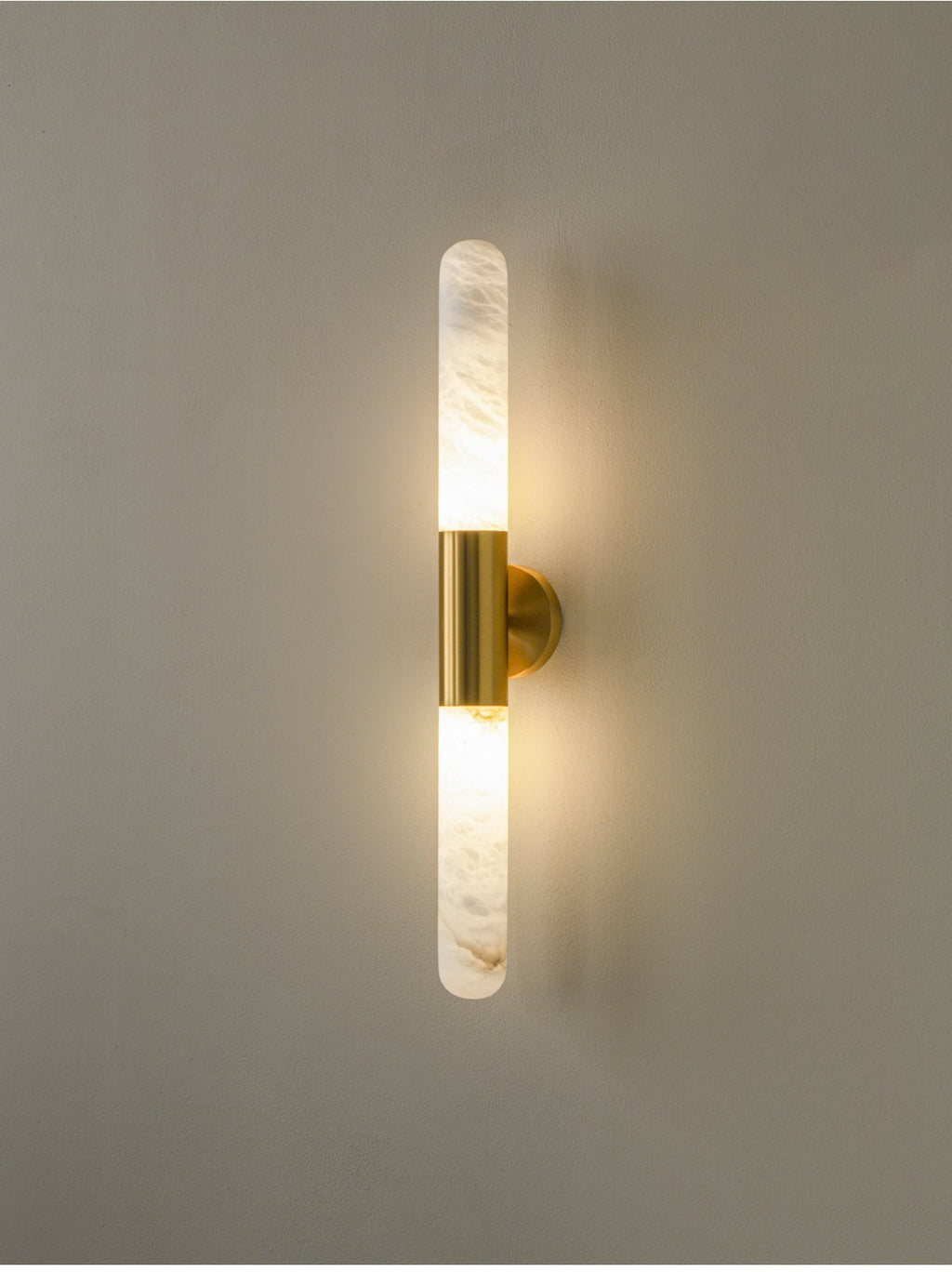 A lit Clérachic gold-and-white wall sconce with soft light glowing around both alabaster ends, on a muted grey-green wall.