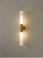 A lit Clérachic gold-and-white wall sconce with soft light glowing around both alabaster ends, on a muted grey-green wall.