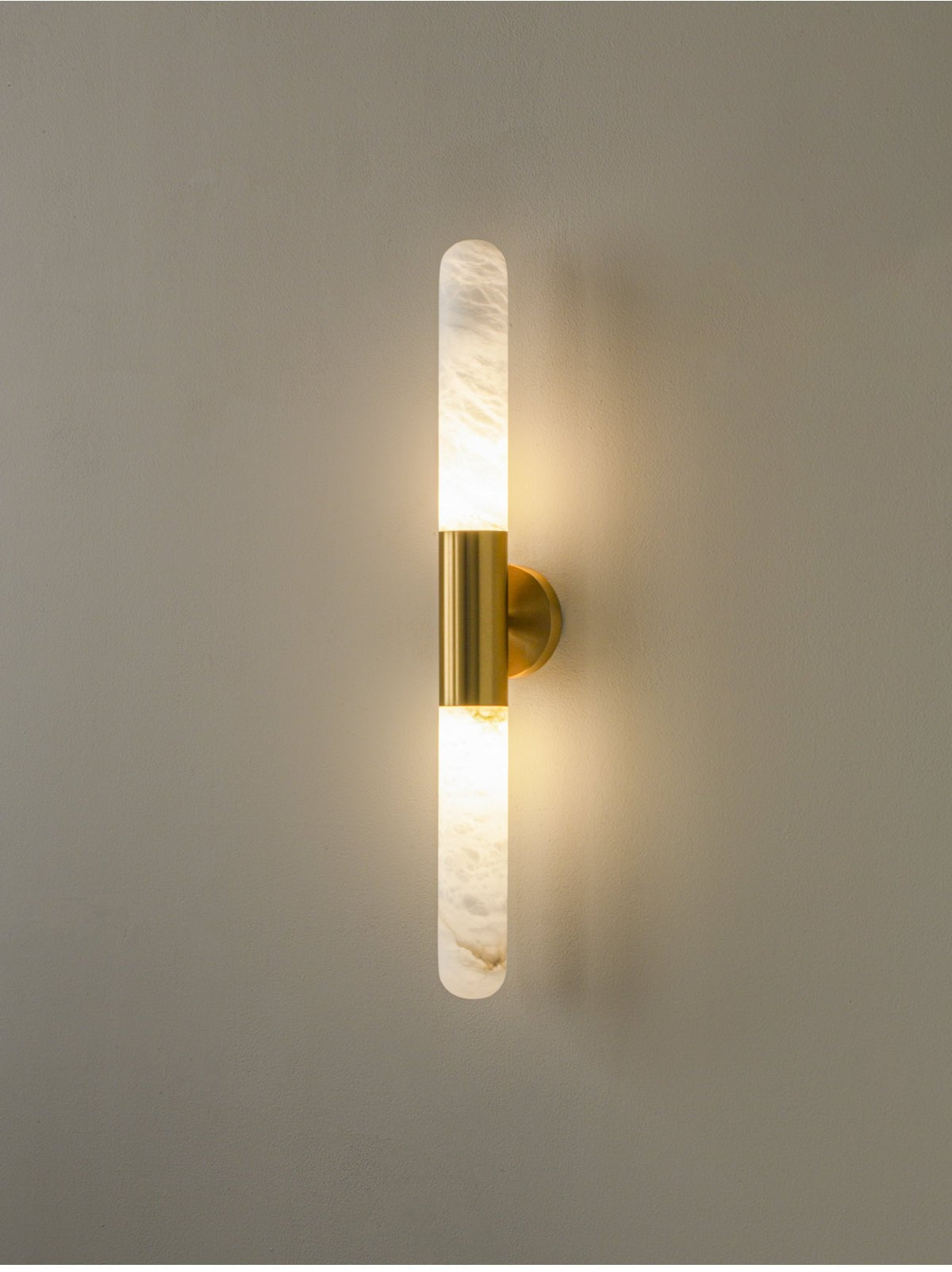 A lit Clérachic gold-and-white wall sconce with soft light glowing around both alabaster ends, on a muted grey-green wall.