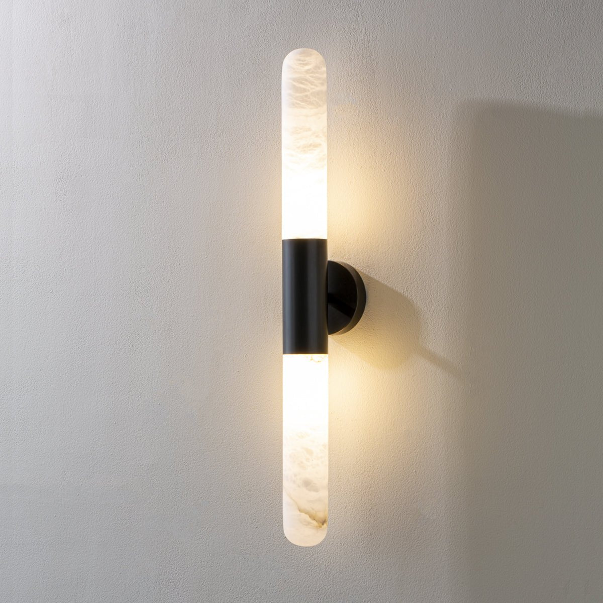 Close, lit view of a black-and-white Clérachic wall sconce on a pale grey wall, with warm light around the alabaster tubes.