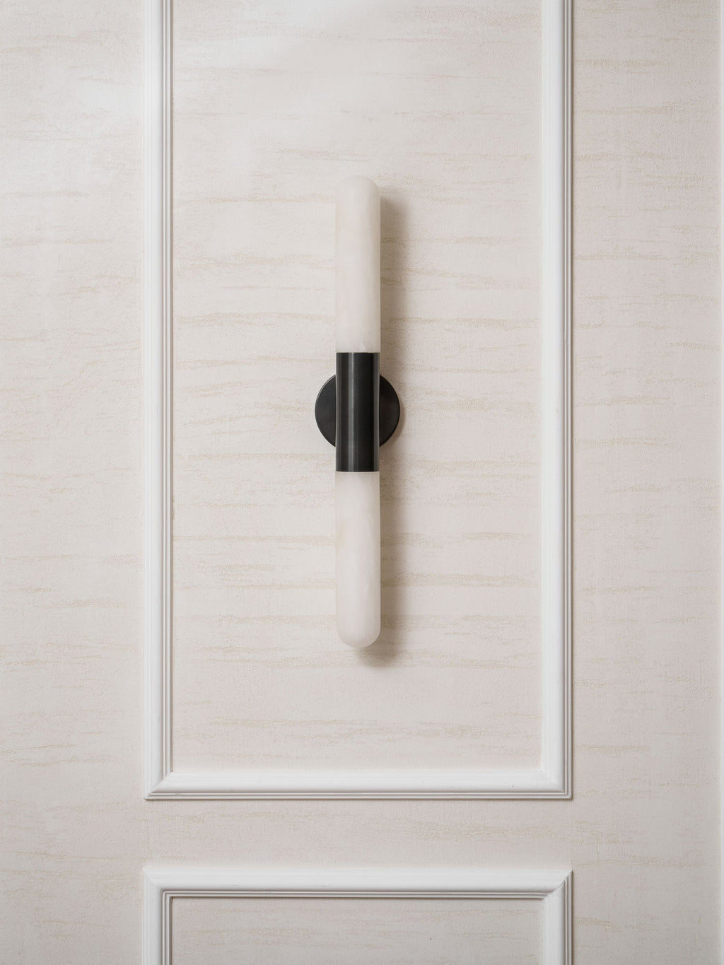 A black-and-white Clérachic wall light on a moulded cream wall panel, unlit, with its alabaster tube catching daylight.