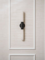 A black-and-white Clérachic wall light on a moulded cream wall panel, unlit, with its alabaster tube catching daylight.