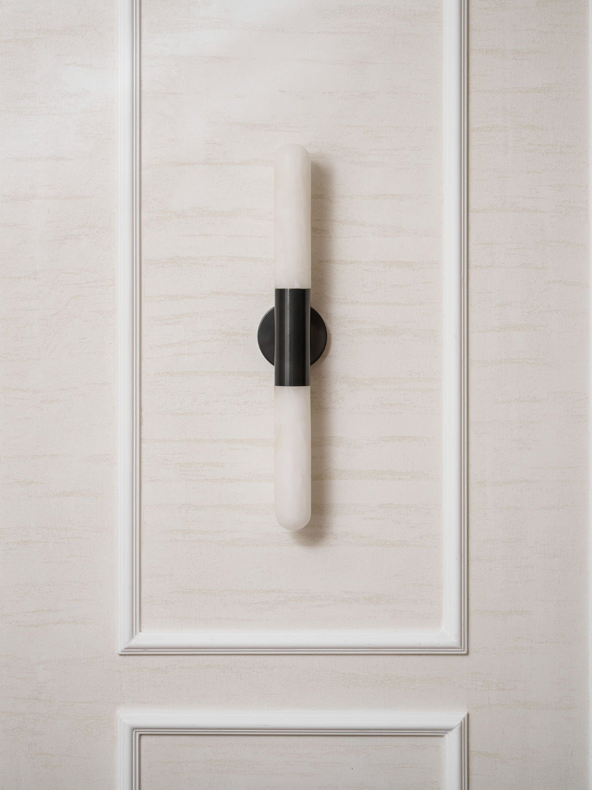 A black-and-white Clérachic wall light on a moulded cream wall panel, unlit, with its alabaster tube catching daylight.