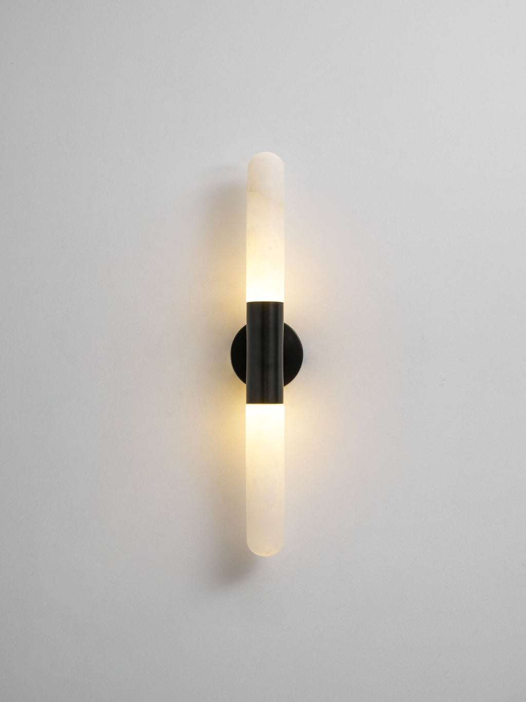 A lit Clérachic black-and-white wall sconce on a light grey wall, with a soft glow around the black band and alabaster ends.