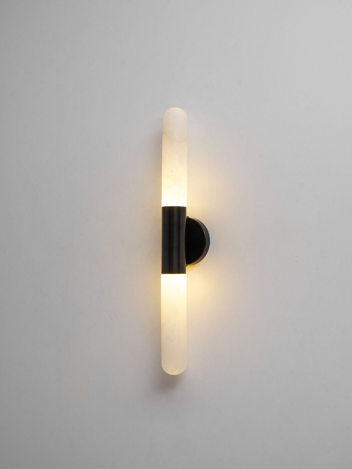 Another lit view of this Clérachic black-and-white wall light on a grey wall, showing a warm glow around both alabaster tubes.