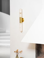 An unlit Clérachic gold-and-white wall sconce on a white wall, above a vase of dried stems and a stone fireplace surround.
