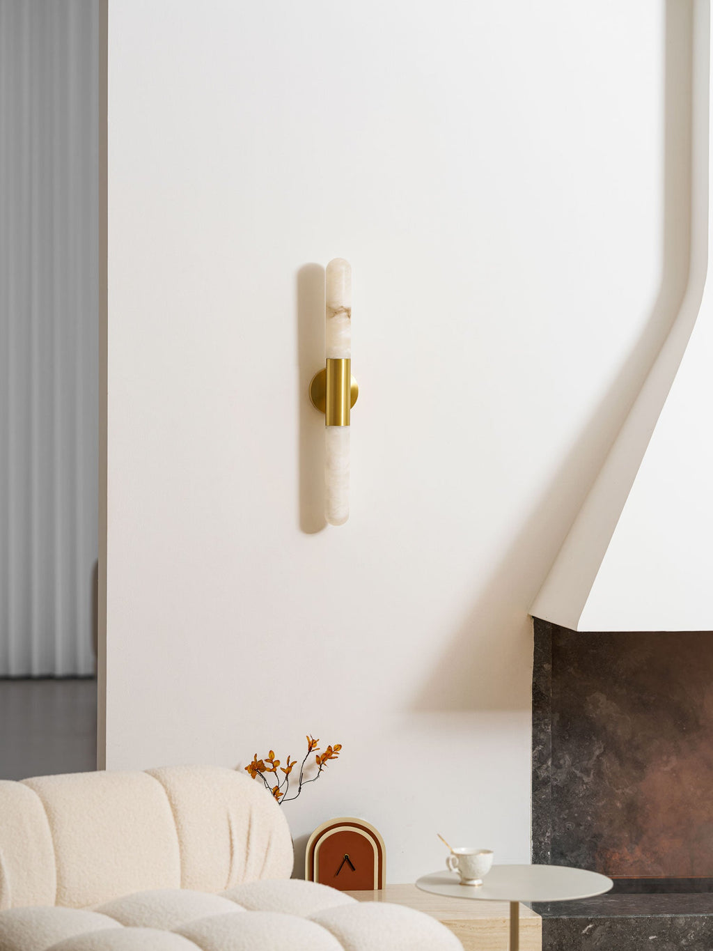 Seen from further back, this Clérachic wall light hangs on a white wall above a boucle sofa, a vase and a stone fireplace.