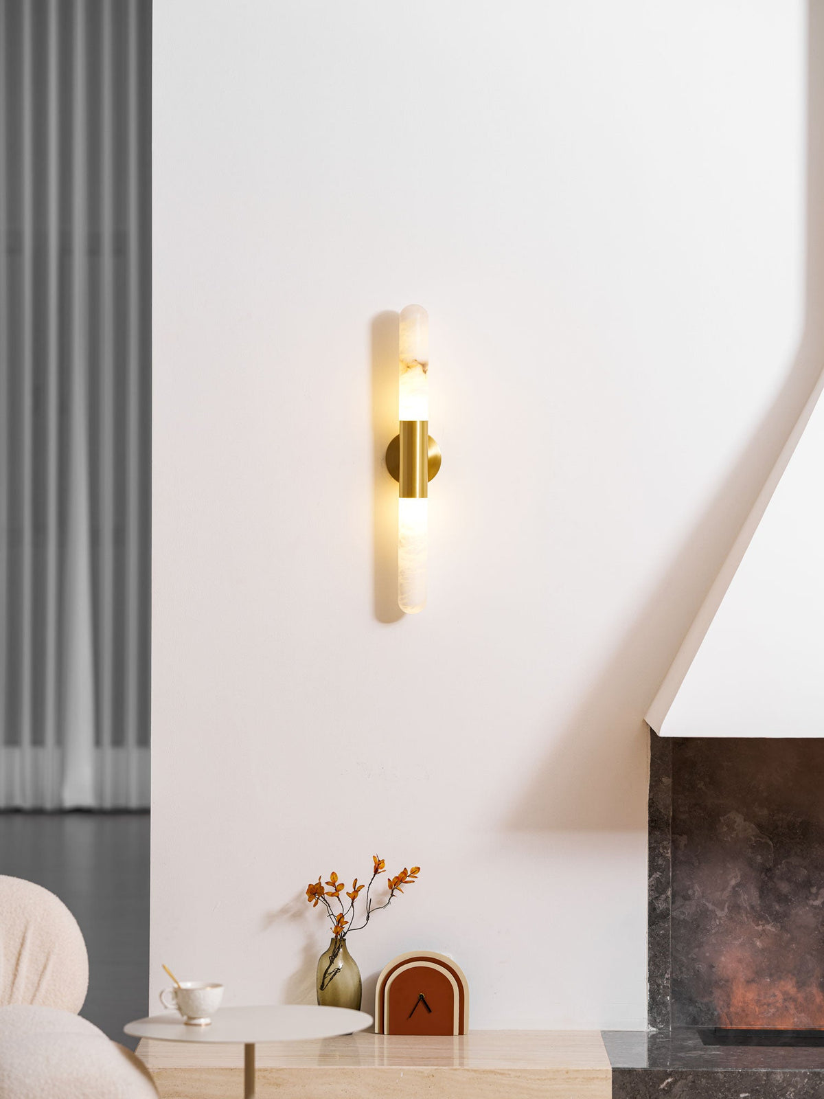 A lit Clérachic gold-and-white wall sconce on a white wall, with a warm glow at the centre and a vase of orange stems below.
