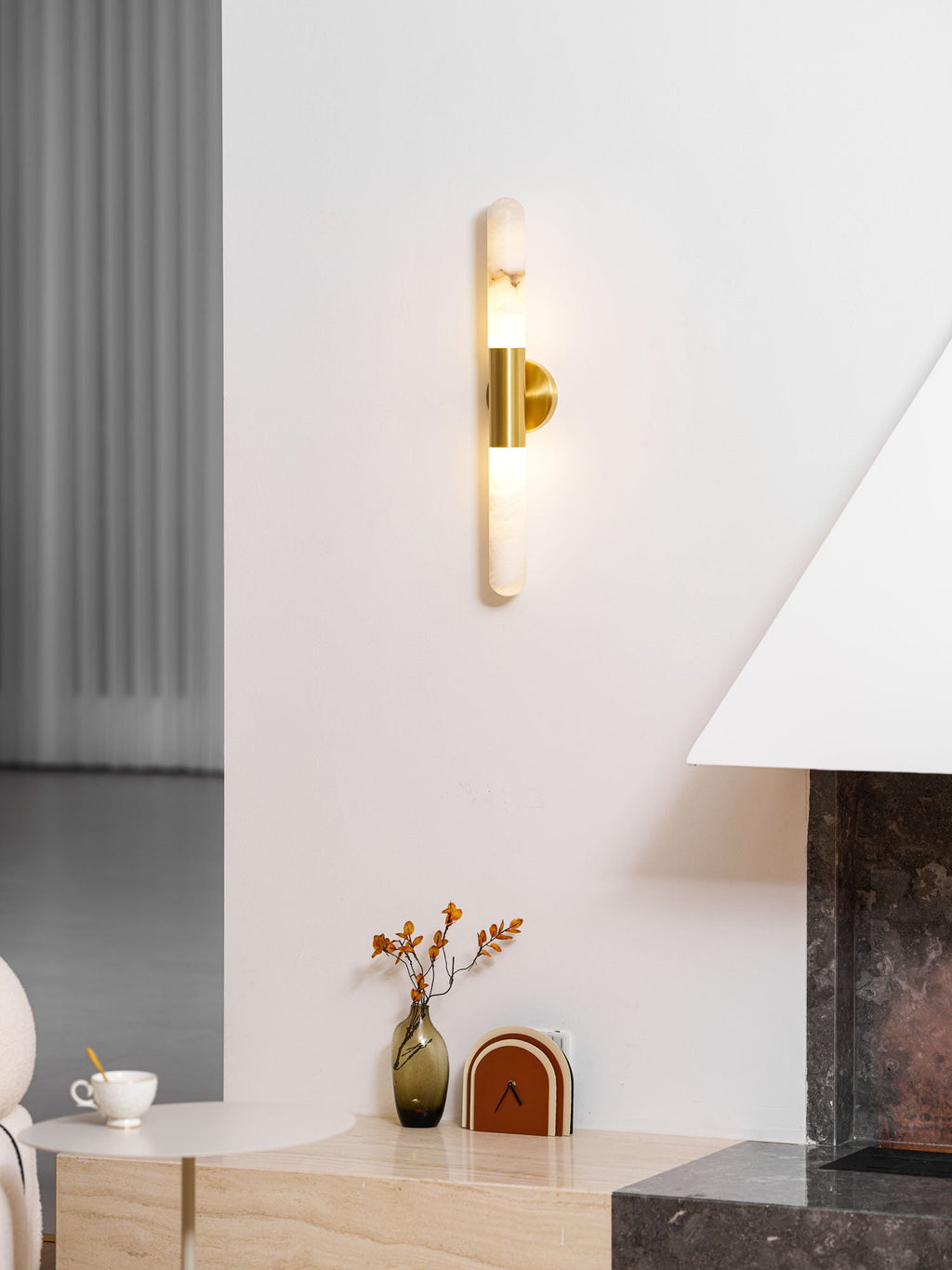 Distant, lit view of this Clérachic gold-and-white wall light on a white wall, with a stone fireplace and a small vase nearby.
