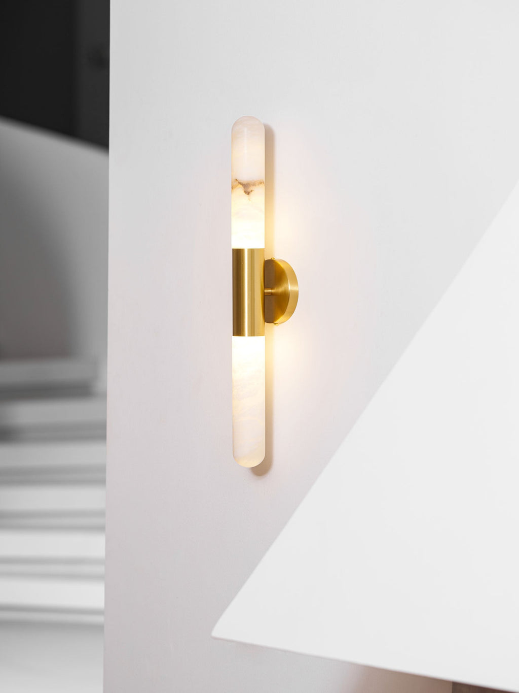 Lit Clérachic wall light with a brass band, close up on a white wall beside the sharp edge of a fireplace hood.