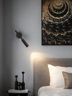 A black Clérachic wall light angled downward above a grey headboard, with a pool of warm light on the wall beside a framed print.