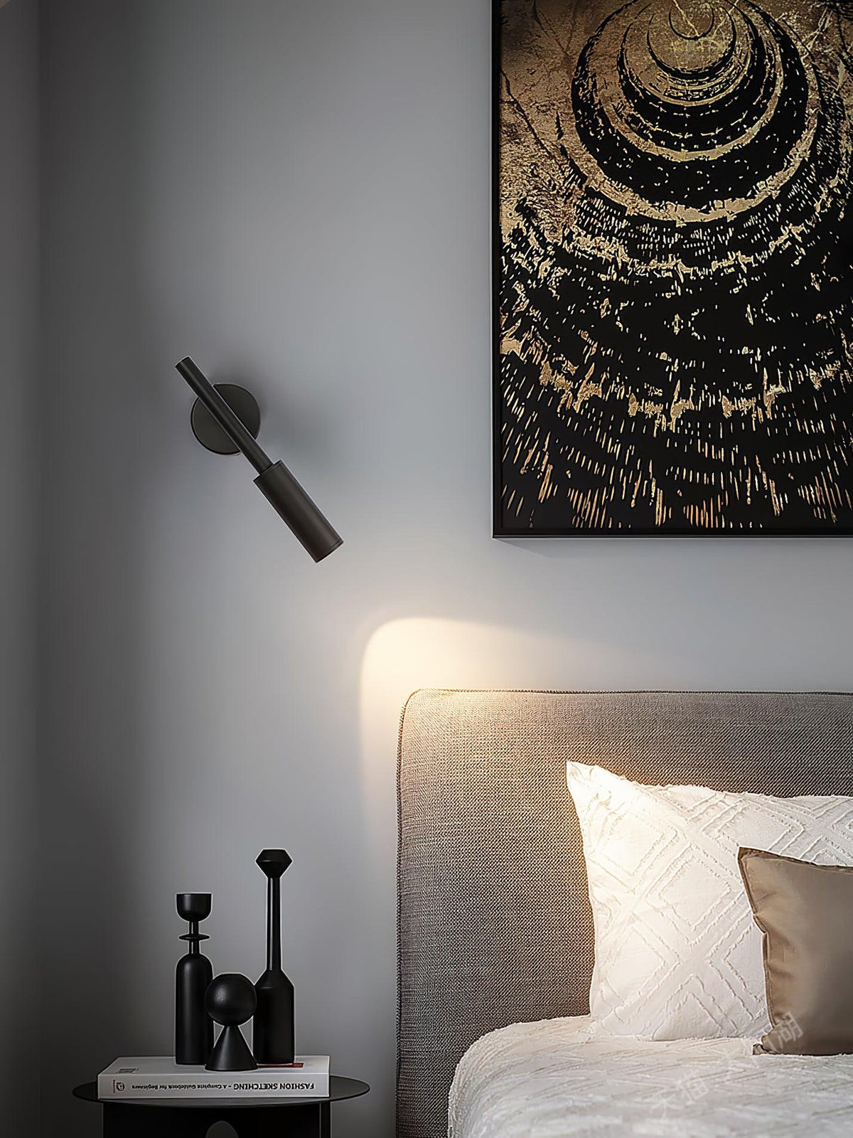 A black Clérachic wall light angled downward above a grey headboard, with a pool of warm light on the wall beside a framed print.