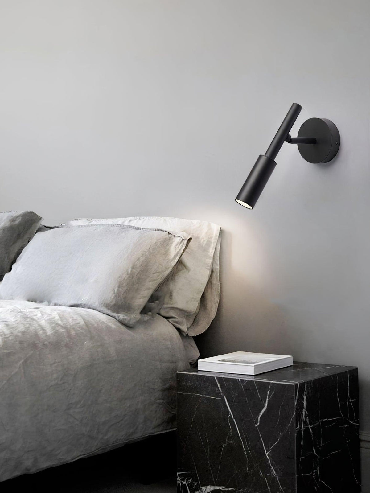 Slim black Clérachic wall light above a grey bed, its downward beam lighting the pillows and a black marble bedside table.