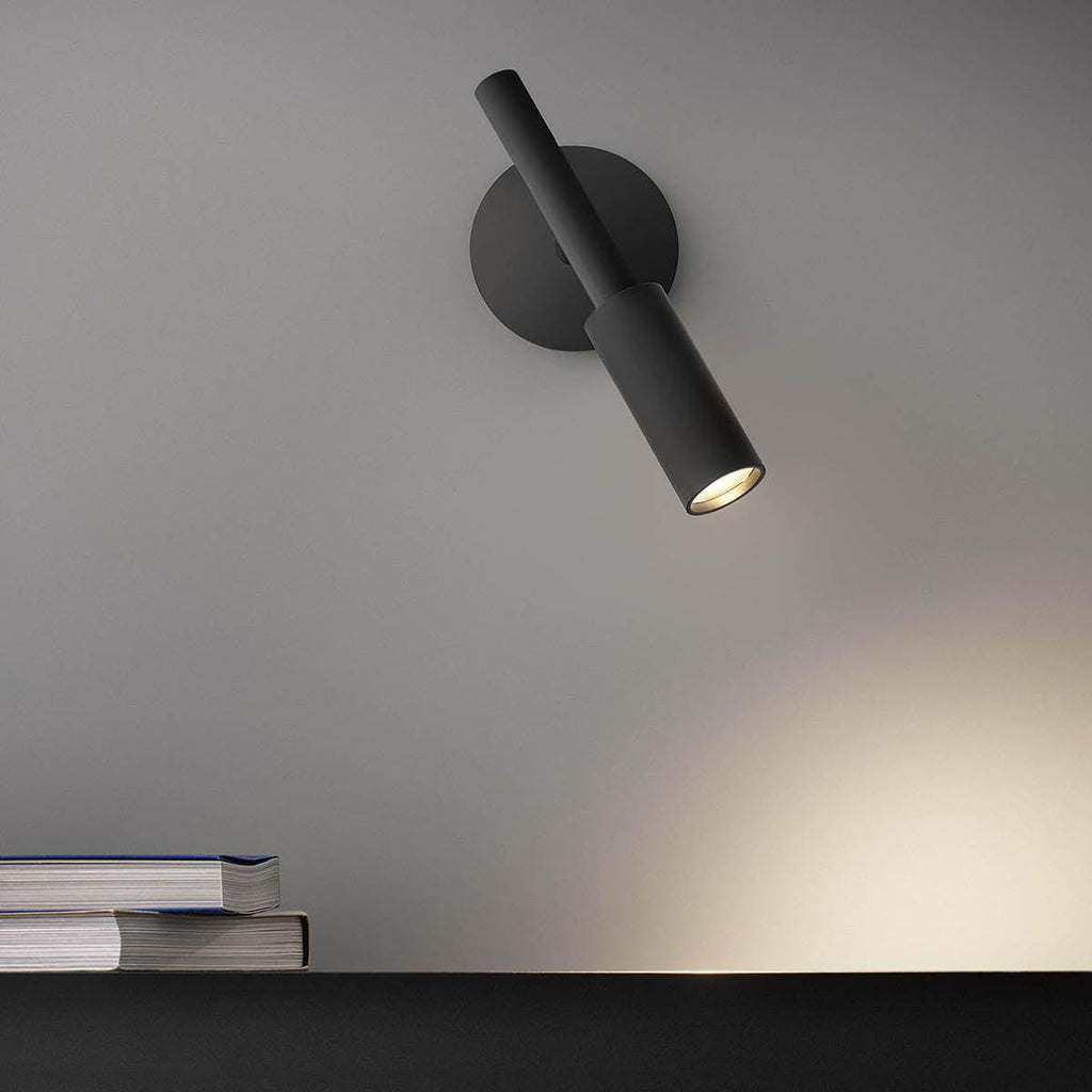 Black Clérachic wall light tilted toward the shelf below, its warm beam falling across the grey wall and a stack of books.